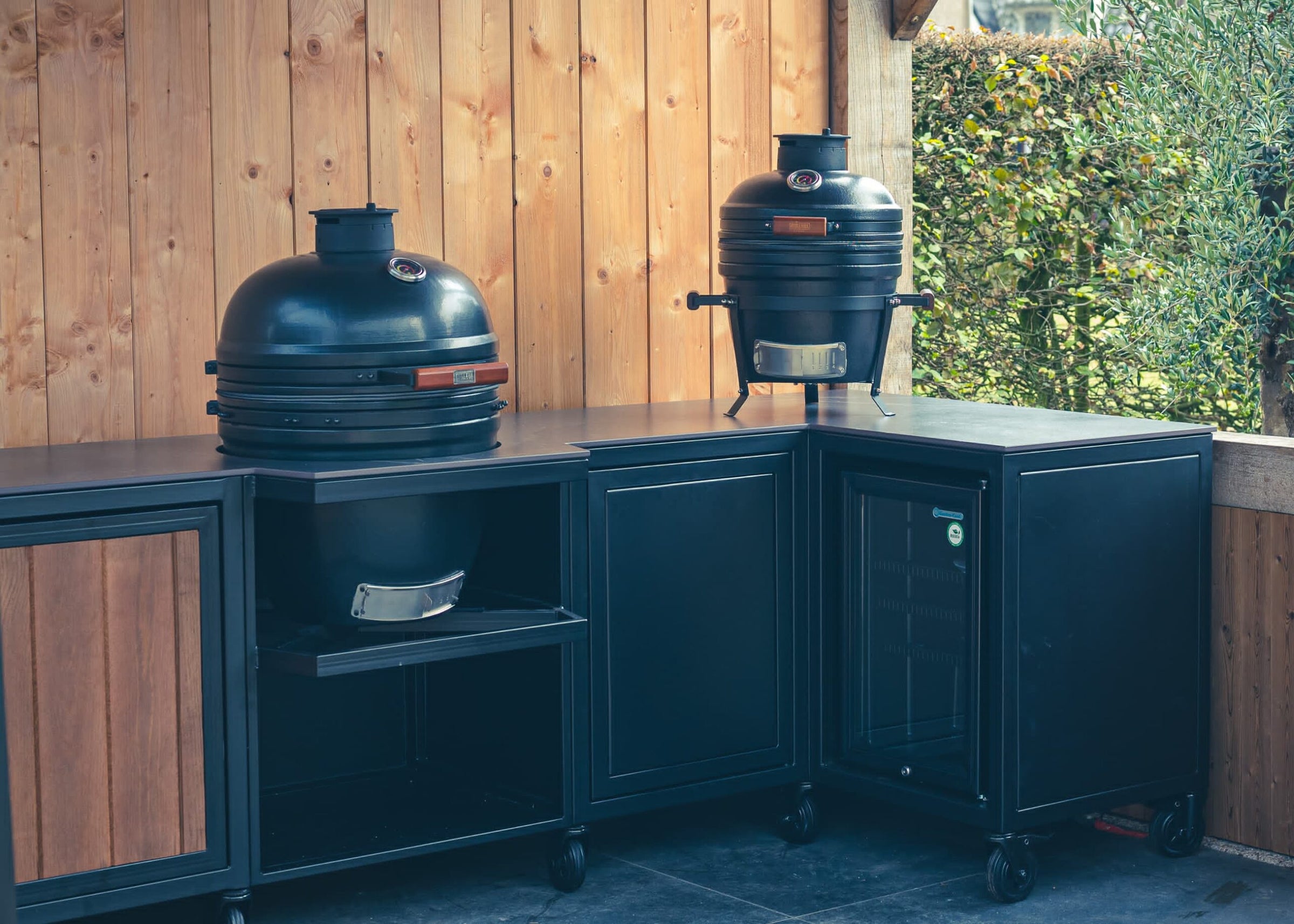 Luxe maatwerk hoek-buitenkeuken met zwarte kasten, ingebouwde koelkast en twee Kamado BBQ’s. Stijlvol, duurzaam en ideaal voor buiten koken